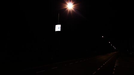 Terschuur, weg in de avond met lantaarnpalen. Aan 1 lantaarnpaal hangt een bord met verlichte reclame. Daar richt je oog zich op